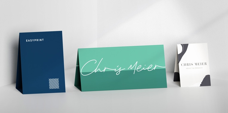 Drei aufgestellte Tischaufsteller in unterschiedlichen Designs: Blau mit Text, türkis mit Handschrift, und weiß mit geometrischem Muster, auf einer hellen Oberfläche.