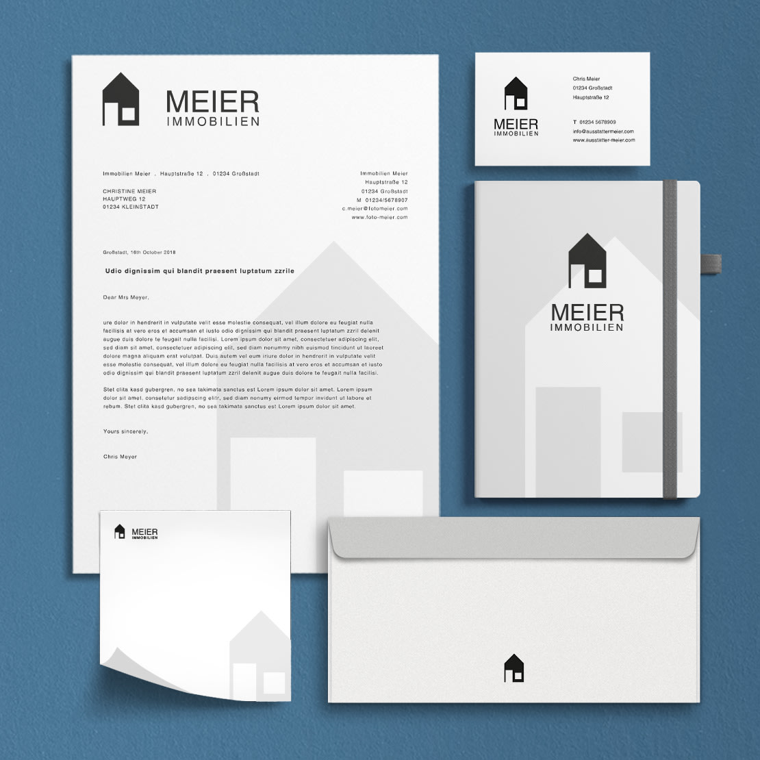 Skrivesaker for eiendomsvirksomhet på blå bakgrunn, inkludert brevpapir, visittkort, notatbok og konvolutt, alt med husets logo og svart tekst.