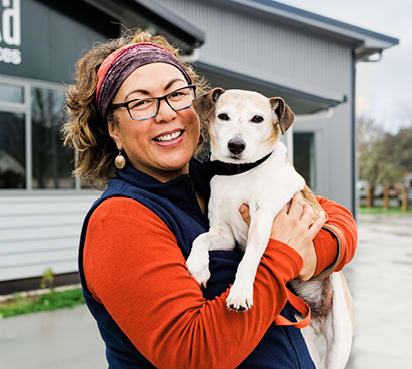 A woman wearing glasses smiles as she holds up a small dog.