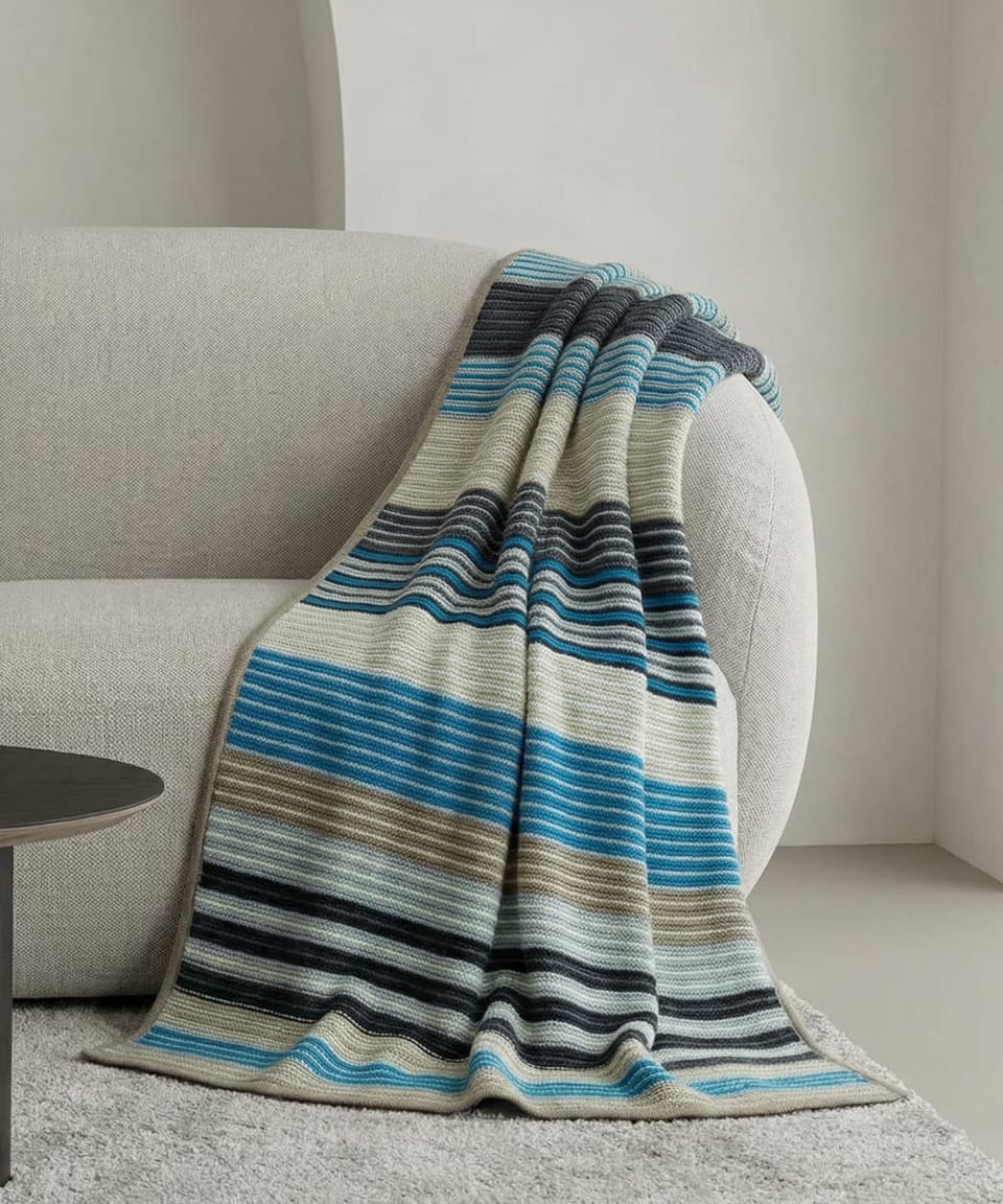 Multi-coloured striped heavyweight cashmere throw in blue, navy, cream and taupe with taupe bound edge, draped over a pale linen round-backed sofa