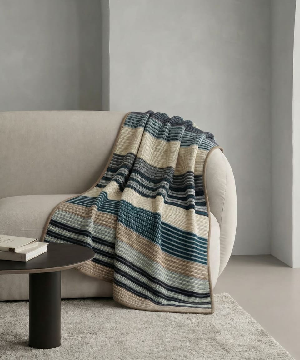 Multi-coloured striped heavyweight cashmere throw in navy, teal, charcoal, cream and taupe with taupe bound edge, draped over a pale linen curved sofa beside a dark round coffee table