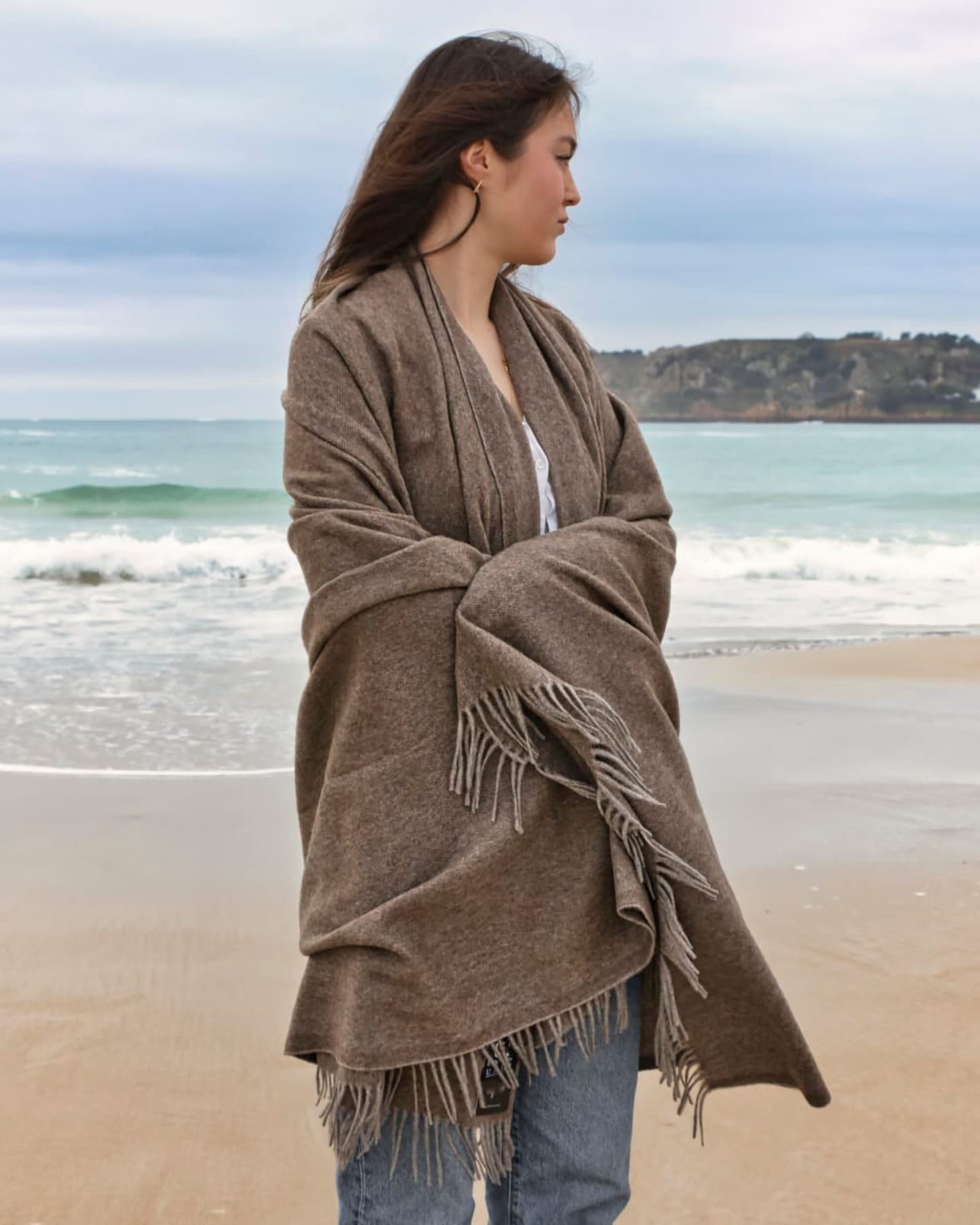 Smiling blonde woman wrapped in olive-taupe yak wool throw over a brown beanie, showing the throw's generous shawl-sized drape