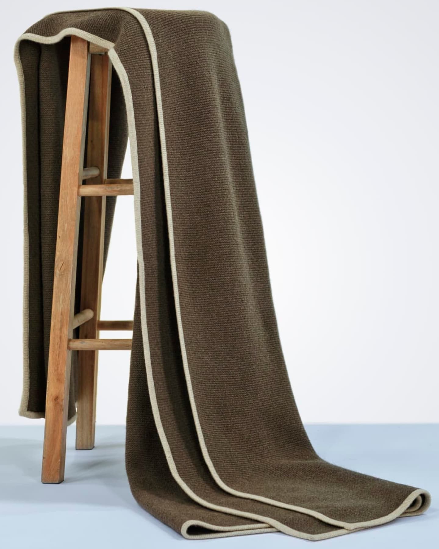 Olive-taupe knitted yak wool throw with cream border draped full-length over a wooden ladder against a white studio background