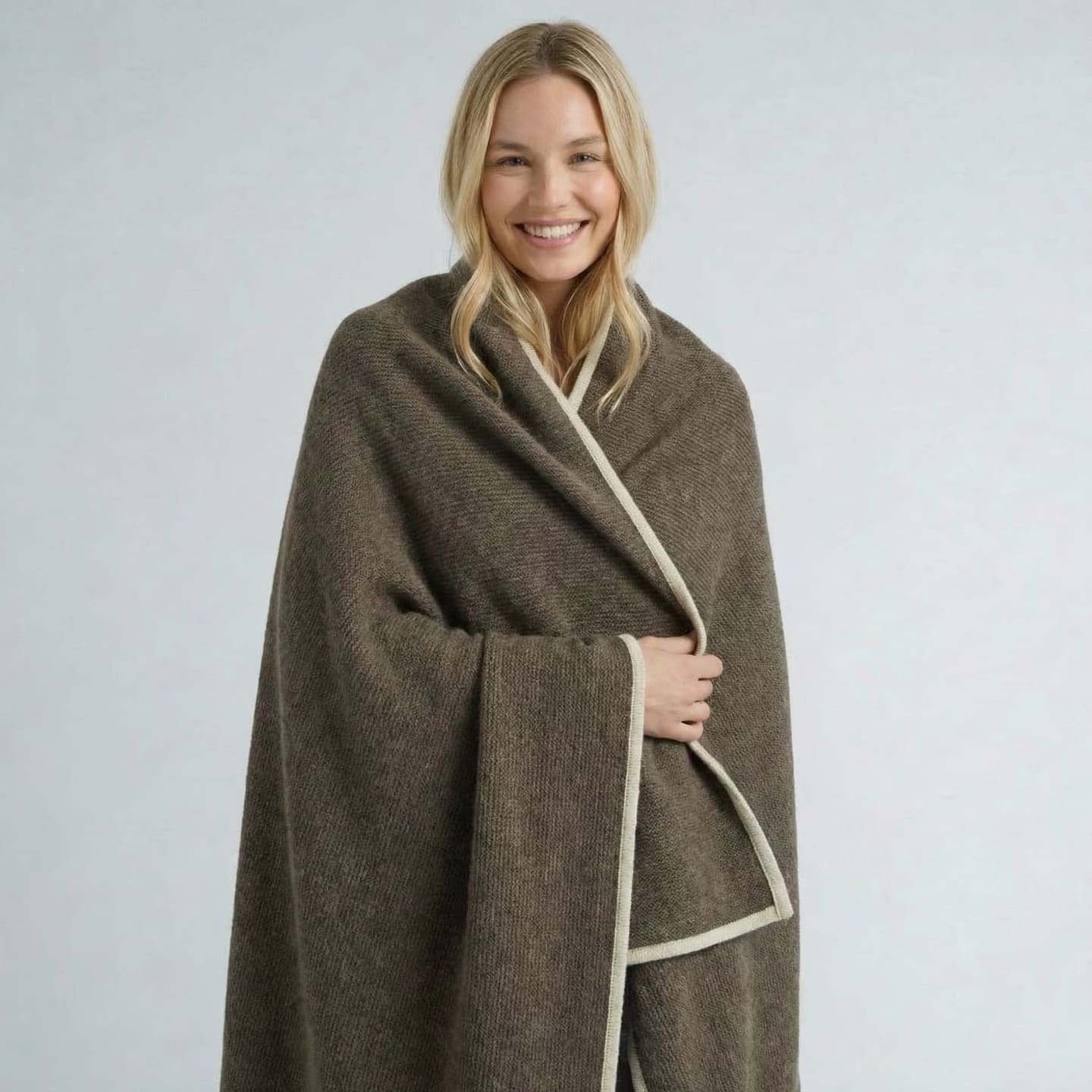 Smiling blonde woman wrapped in an olive-taupe knitted yak wool throw with cream border, showing the throw's generous wrap-sized drape