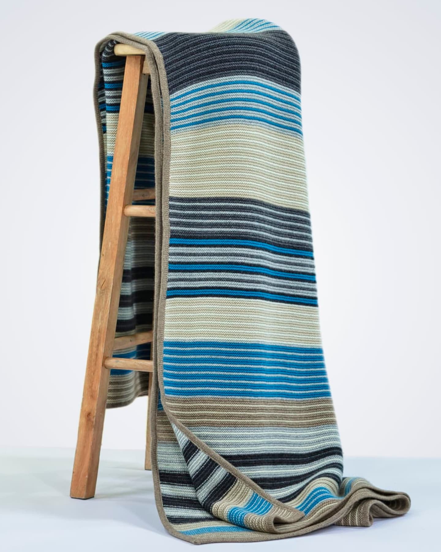 Multi-coloured striped heavyweight cashmere throw in blue, navy, charcoal, cream and taupe draped full-length over a wooden ladder against a white studio background