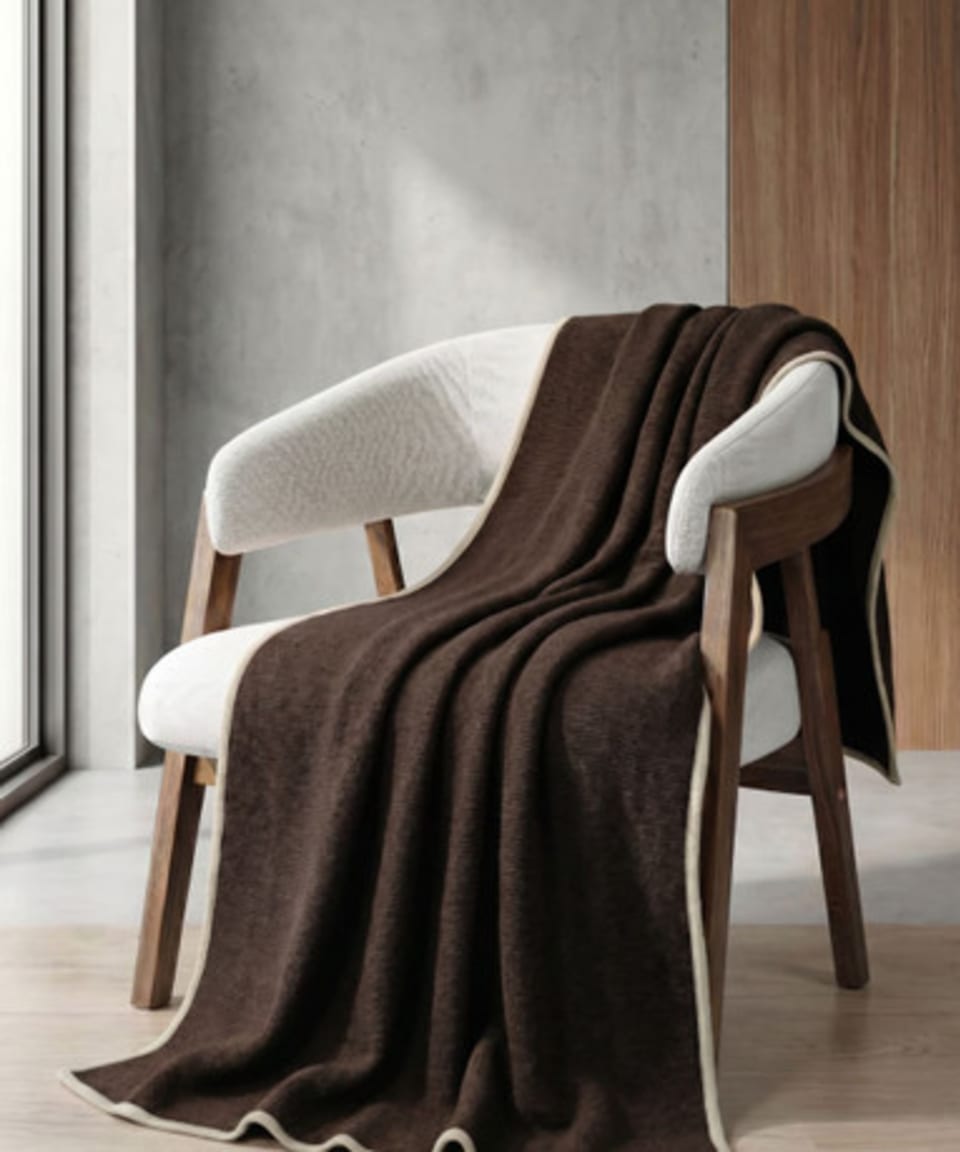 Dark chocolate brown knitted yak wool throw with cream border draped over a white-upholstered walnut armchair in a concrete-and-walnut interior