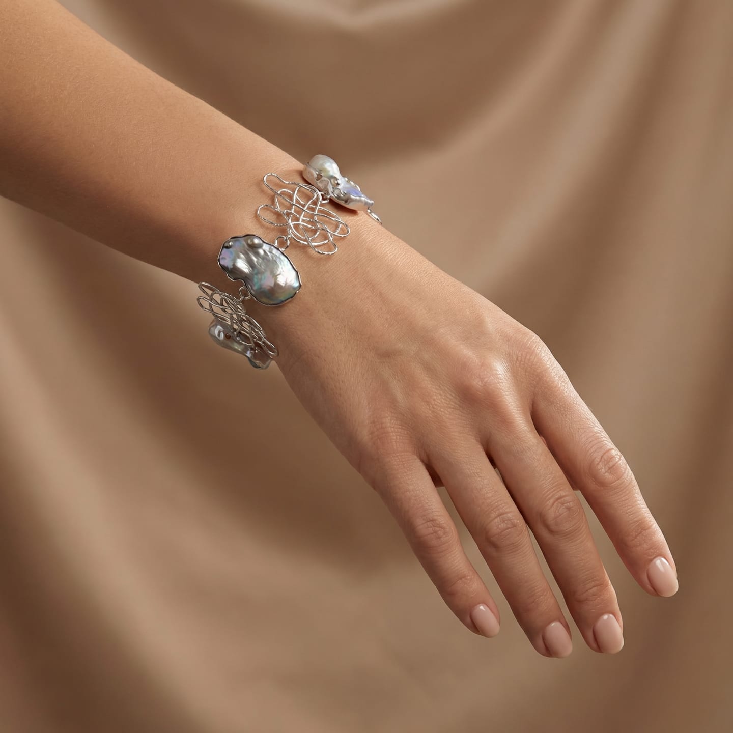 Model wearing the Signature Dark Keshi Pearl Chain Bracelet on her wrist, large flat peacock-grey keshi pearls alternating with openwork silver wire-scribble charms, against draped beige silk.