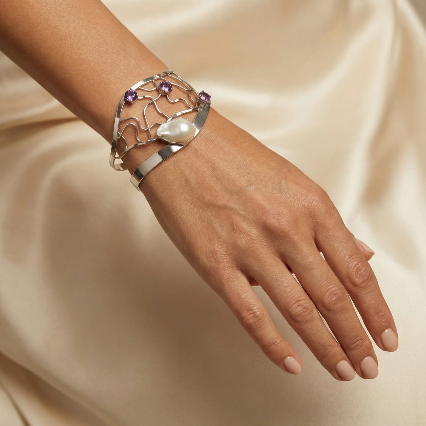 Open silver lattice cuff bracelet with branching wires, set with a large baroque pearl and three round amethysts, worn on the wrist against ivory satin