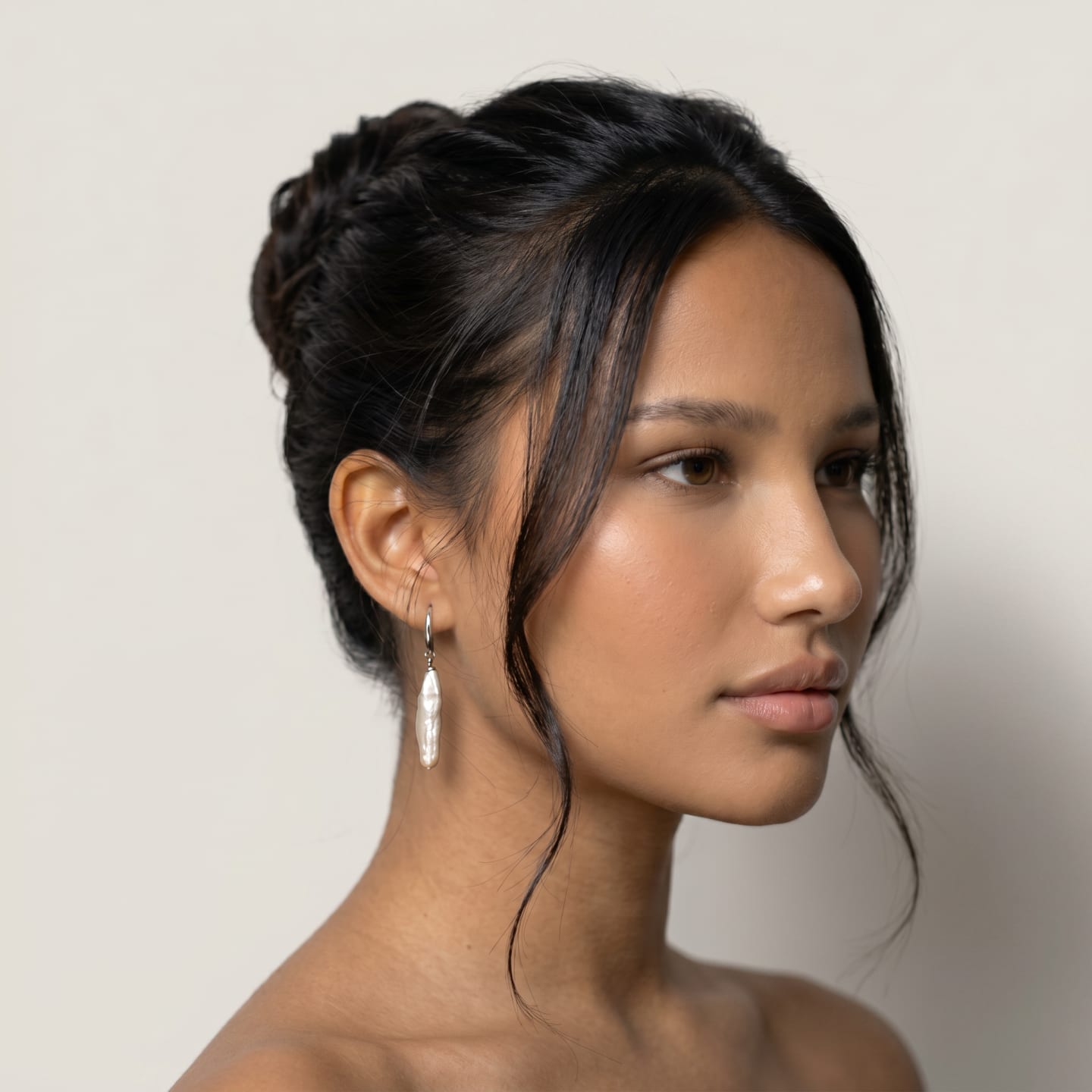 Model wearing the Classic Elongated Peach Pearl Drop Earrings, side-profile portrait against a soft cream backdrop, the right earring shown clearly: a single elongated biwa pearl drop suspended from a silver shepherd's-hook ear wire that curves through the earlobe.