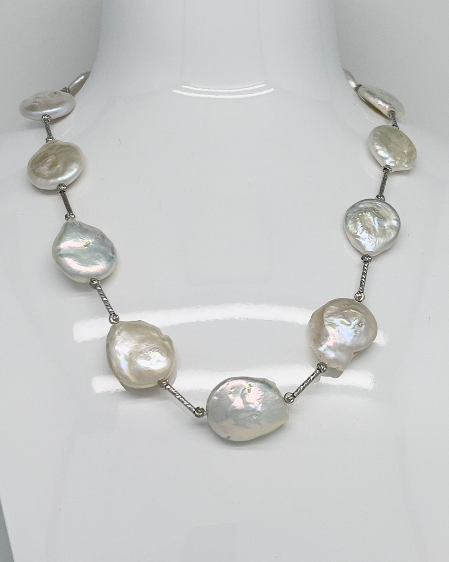 Close-up of the necklace draped on a white bust, white coin pearls with strong rainbow iridescence linked by twisted rope-textured silver bar segments