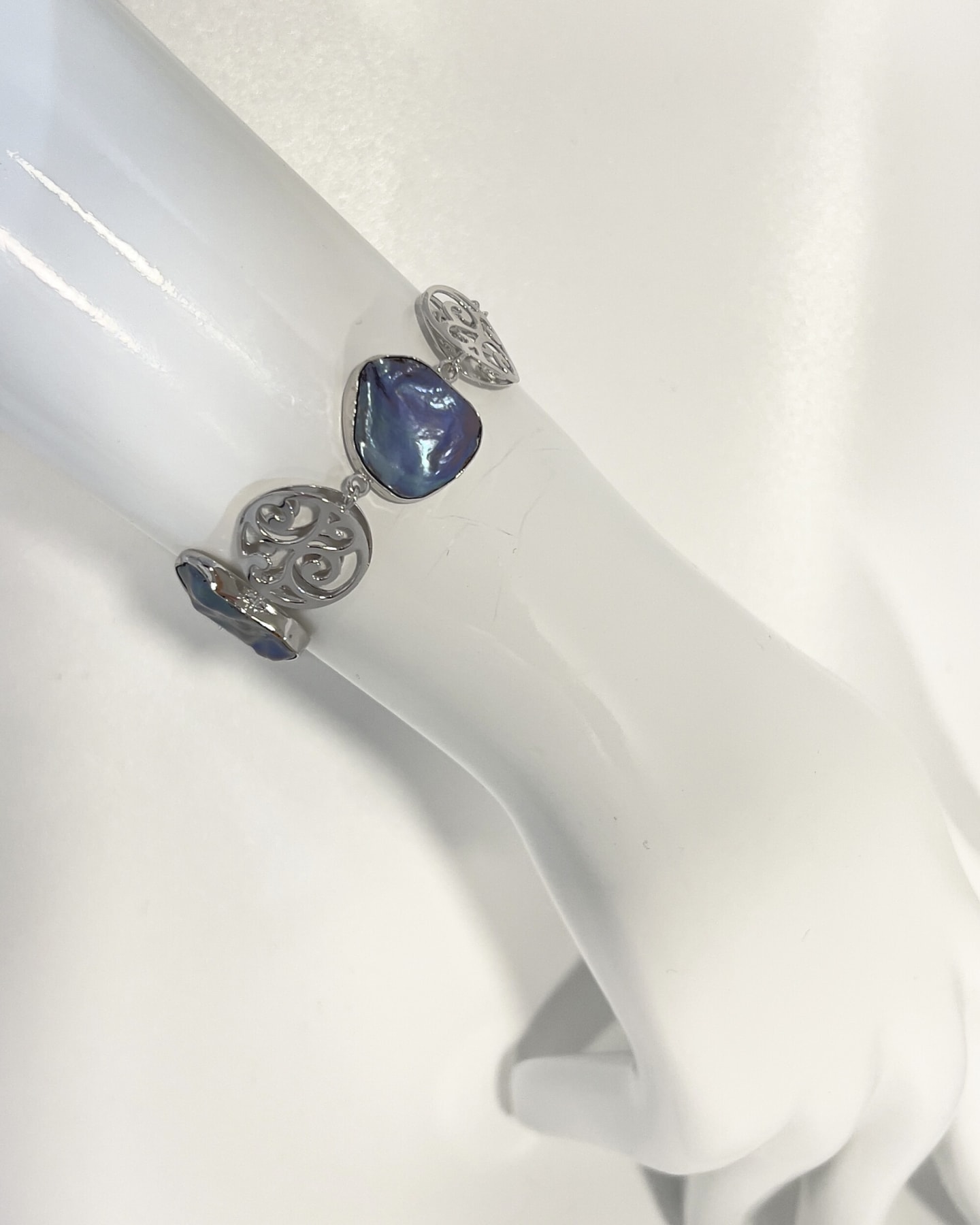 Close-up of a silver filigree scrollwork medallion flanked by two bezel-set dark keshi pearls, on a white display rod.