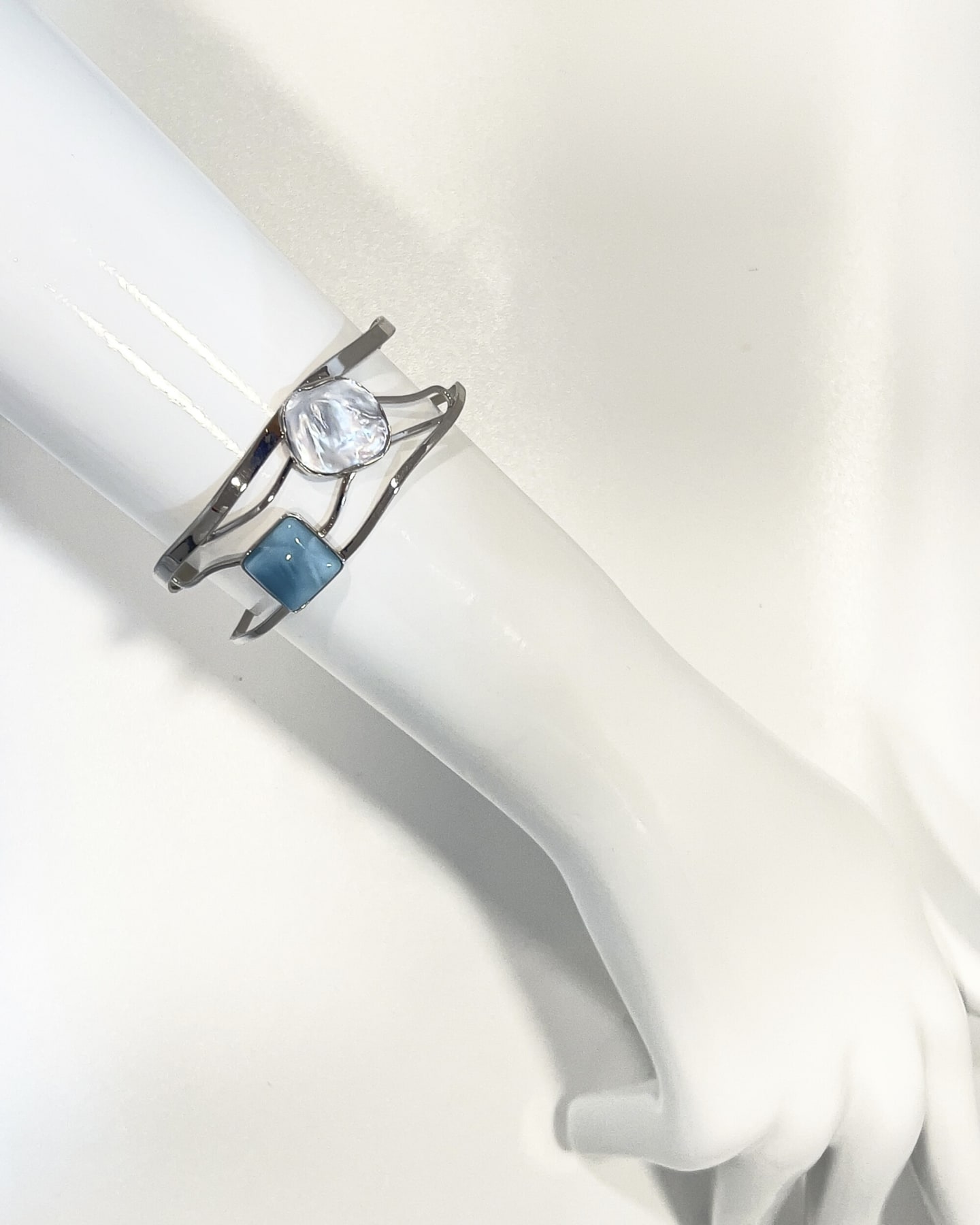 Polished silver-toned cuff bangle with an asymmetrical lattice band, set with a white iridescent keshi pearl and a square blue larimar stone, shown on a plain white backdrop.