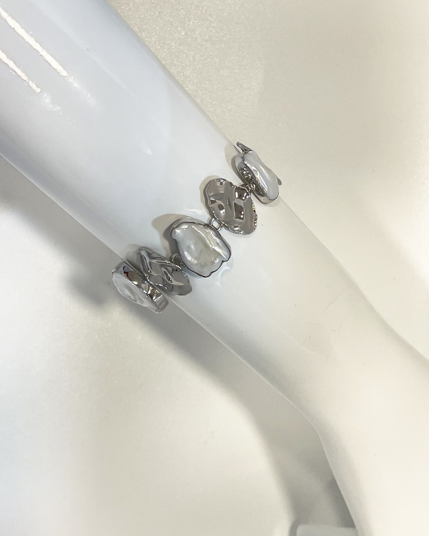 Bracelet draped over a glossy white curved surface on a textured cream background, alternating sculpted silver leaf elements with flat irregular white keshi pearls, soft iridescent pearl lustre and broken reflections across the silver.