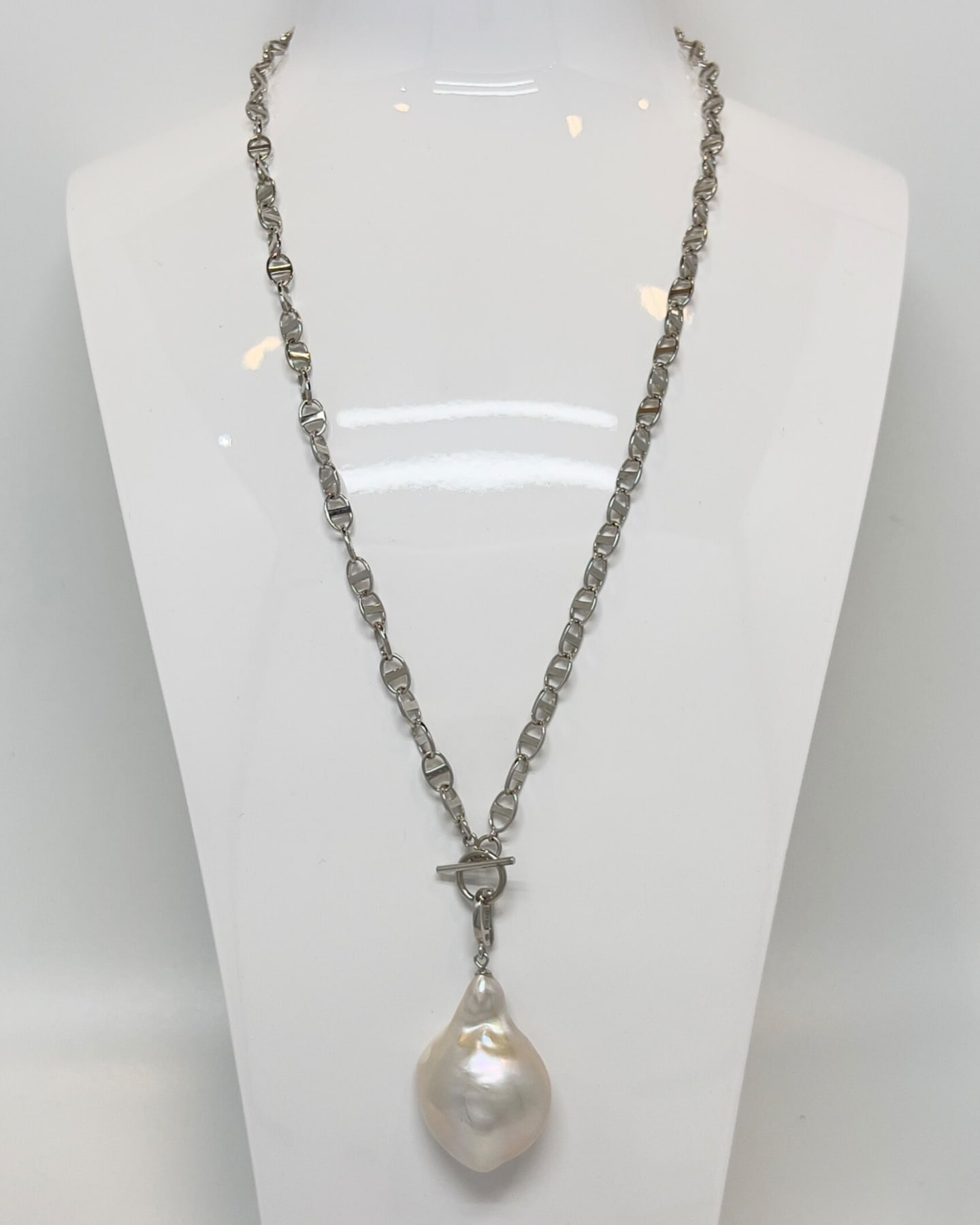 Model wearing the Unur Classic Baroque Pearl Toggle Pendant Necklace, a substantial polished silver cable-link chain with a front-facing toggle clasp at centre, suspending a single large irregular white baroque freshwater pearl drop, paired with a beige openwork knit shawl
