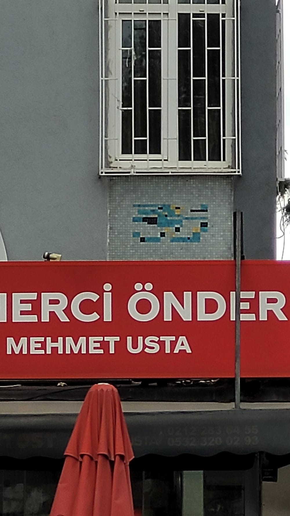 Ferruh Başağa’nın 4 numaralı panosundan geriye kalan küçük mozaik parçası, büyük kırmızı bir dükkân tabelasının hemen üstünde