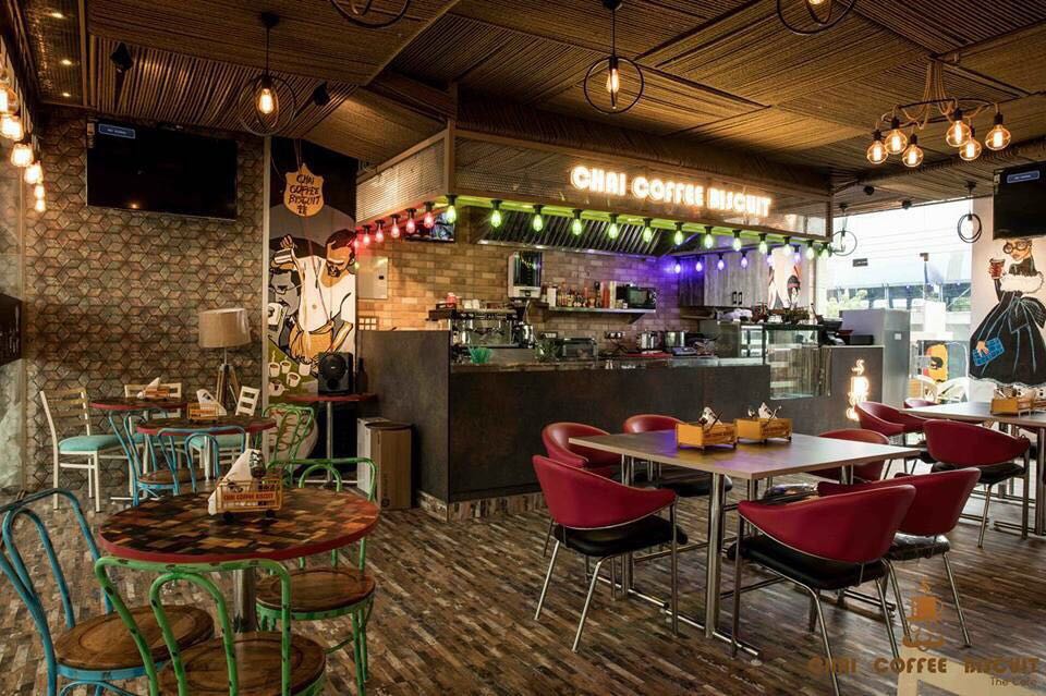Design of a coffee shop with vibrant colored seats and wooden top tables lit by hanging lights by Vikas Swami Contemporary | Interior Design Photos & Ideas