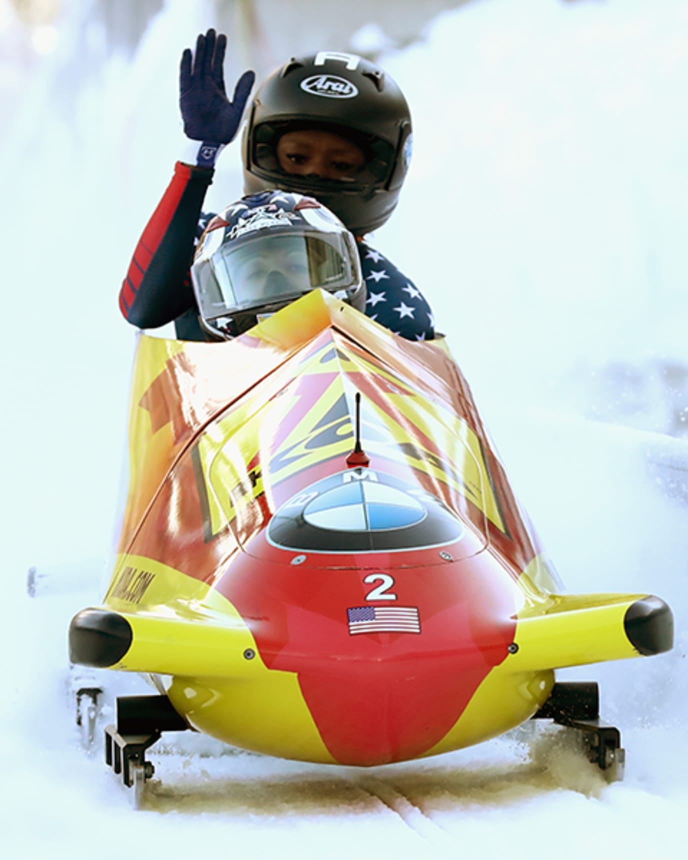 Aja Evans waves in the back of a two-woman red and yellow bobsled while wearing a helmet and red and blue body suit during an event