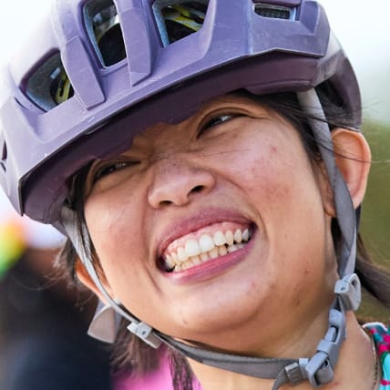 An athlete wearing a helmet and protective gloves raises her hands and smiles in front of the finish line at a race. She is sitting in a racing wheelchair.