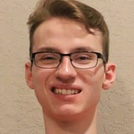 Jenson Van Emburgh smiles and raises both arms, wearing glasses, a red United States short-sleeve shirt, and black pants while sitting in a wheelchair