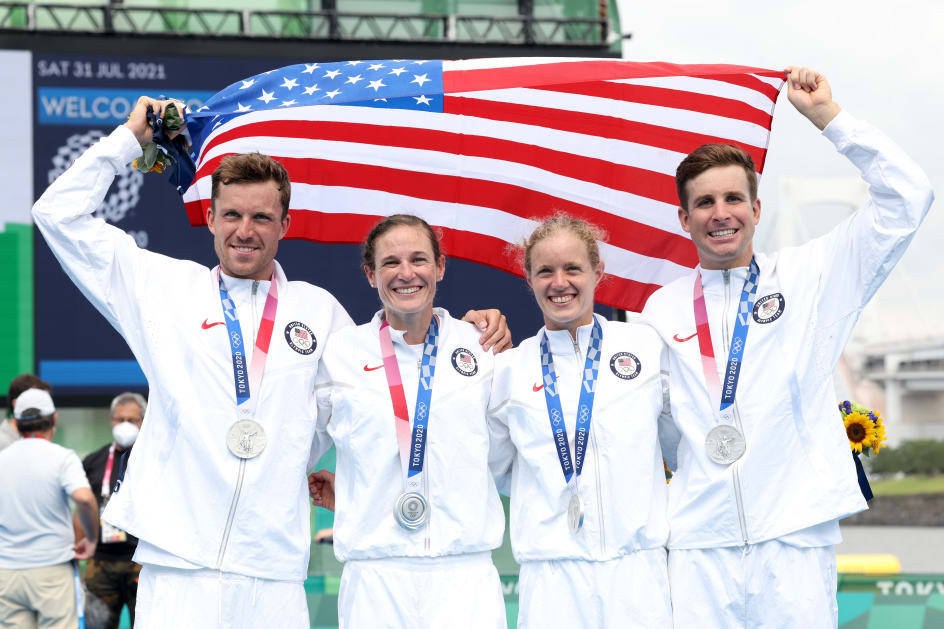 The 2020 U.S. Olympic mixed relay triathlon team - Katie Zaferes, Kevin McDowell, Taylor Knibb, and Morgan Pearson - pose for a photo with silver medals around their necks. Kevin and Morgan are holding onto either side of a U.S. flag, which they're holding up and behind the four of them.