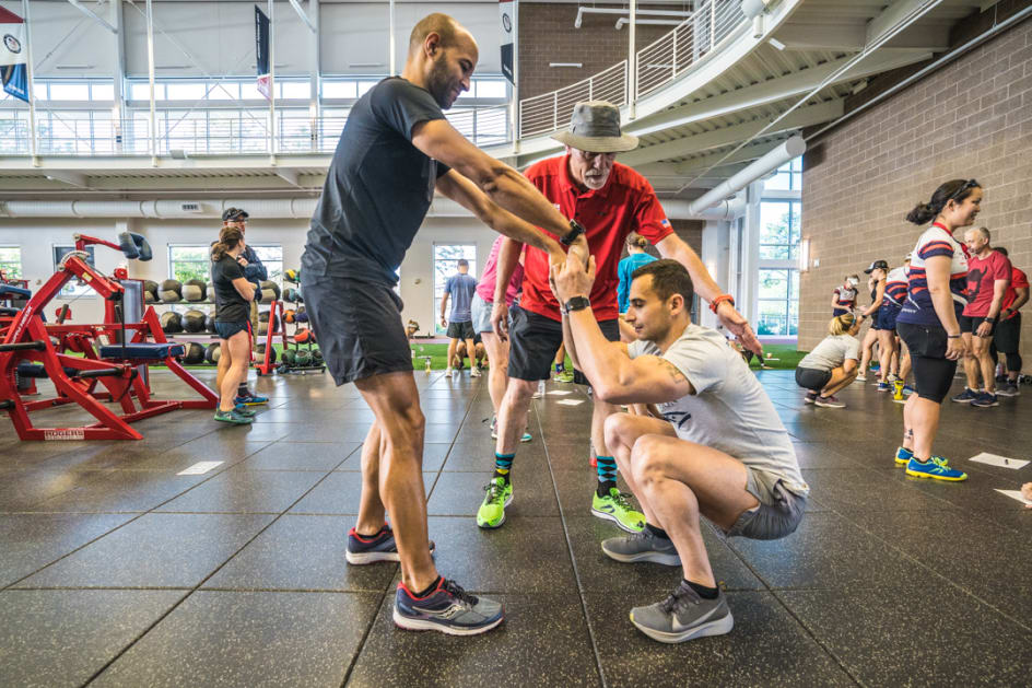 A coach (middle back) guides an athlete's form during a workout that involves hanging onto a partner's hands and squatting.