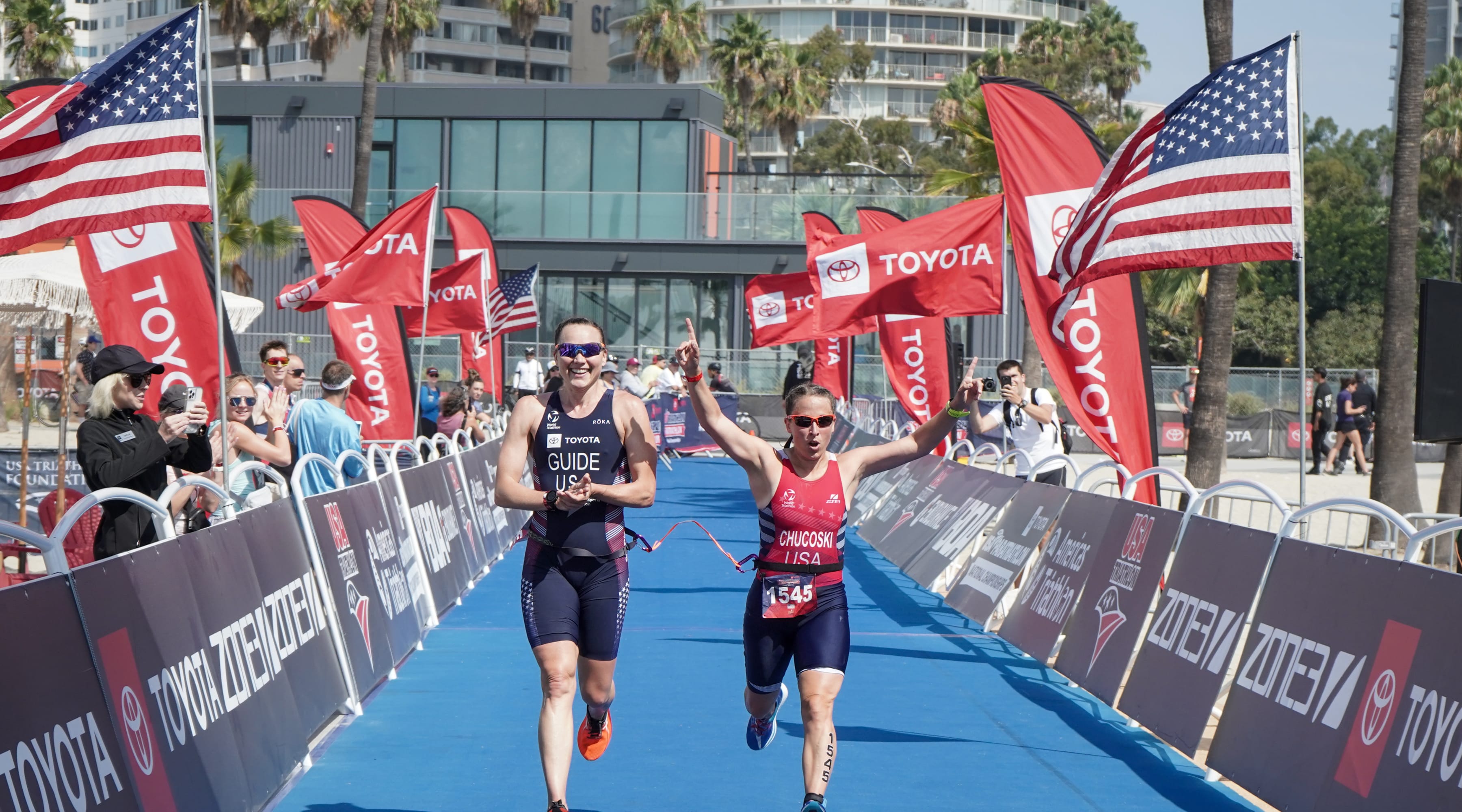 Deb Chuckoski raises her arms while crossing the finish line at a race. Her guide, to whom Deb is tethered, is smiling and clapping her hands. The two athletes are connected by a tether.