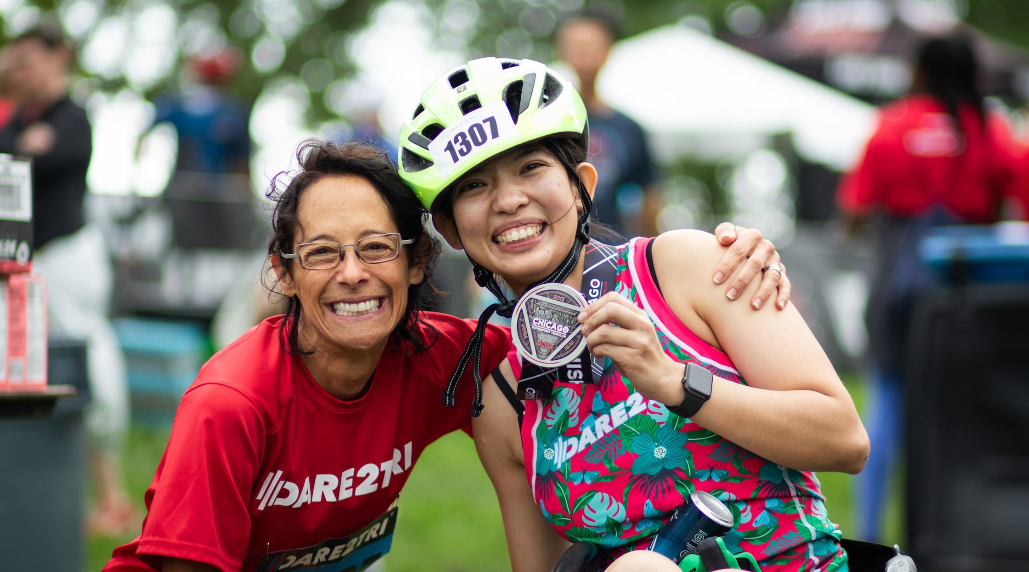 An athlete wearing a helmet with a racing number on it and sitting in a racing wheelchair smiles and holds up a medal. A woman with a bib that says guide crouches down and has her arm around the athlete.