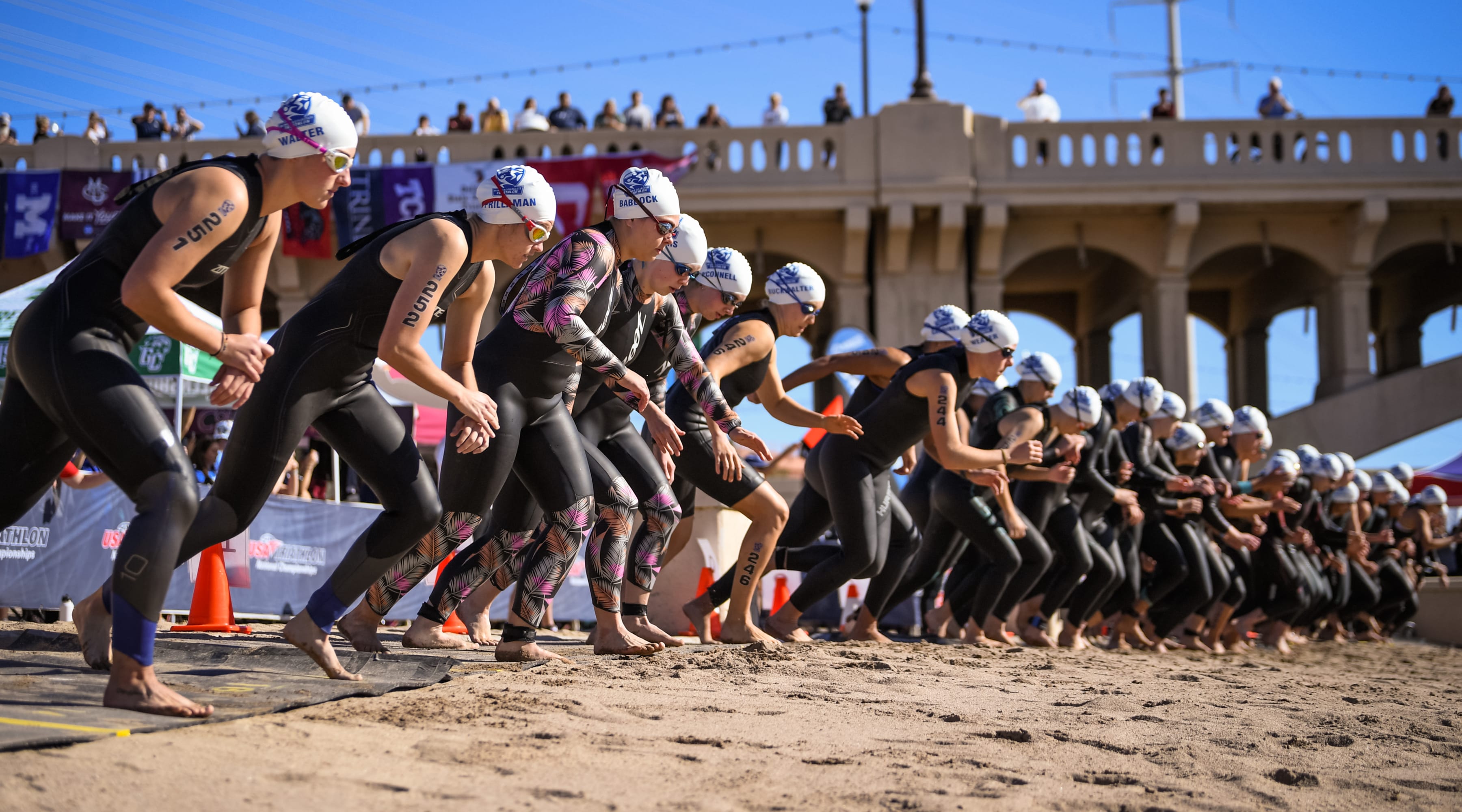 Athletes run on the beach toward water wearing wetsuits and swim caps during a draft-legal race.