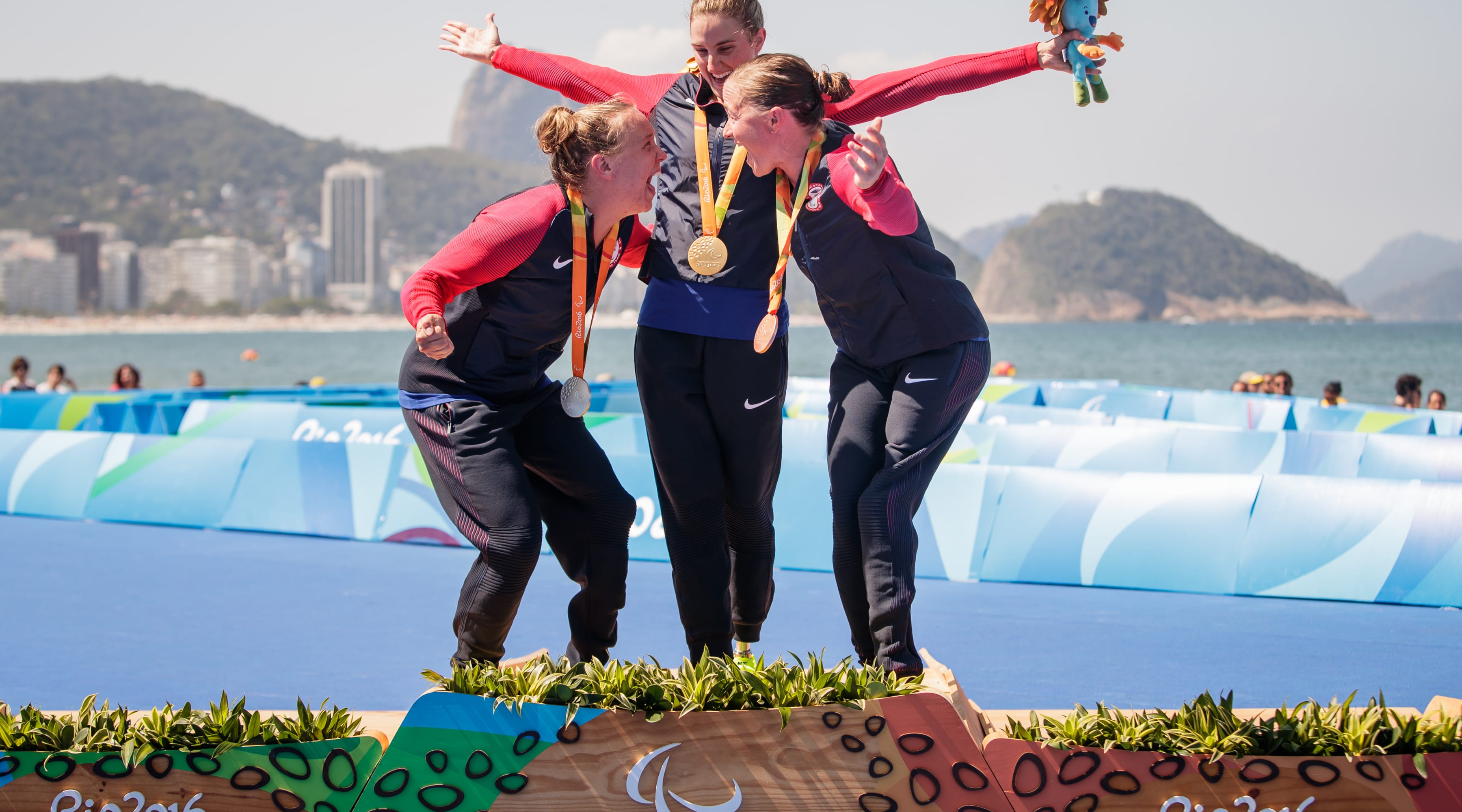 Allysa Seely, Hailey Danz, and Melissa Stockwell celebrate and yell while standing atop the podium at the 2016 Paralympic games. All three have medals around their necks, and Melissa Stockwell (middle) is holding a stuffed toy awarded to gold medalists in her left hand.