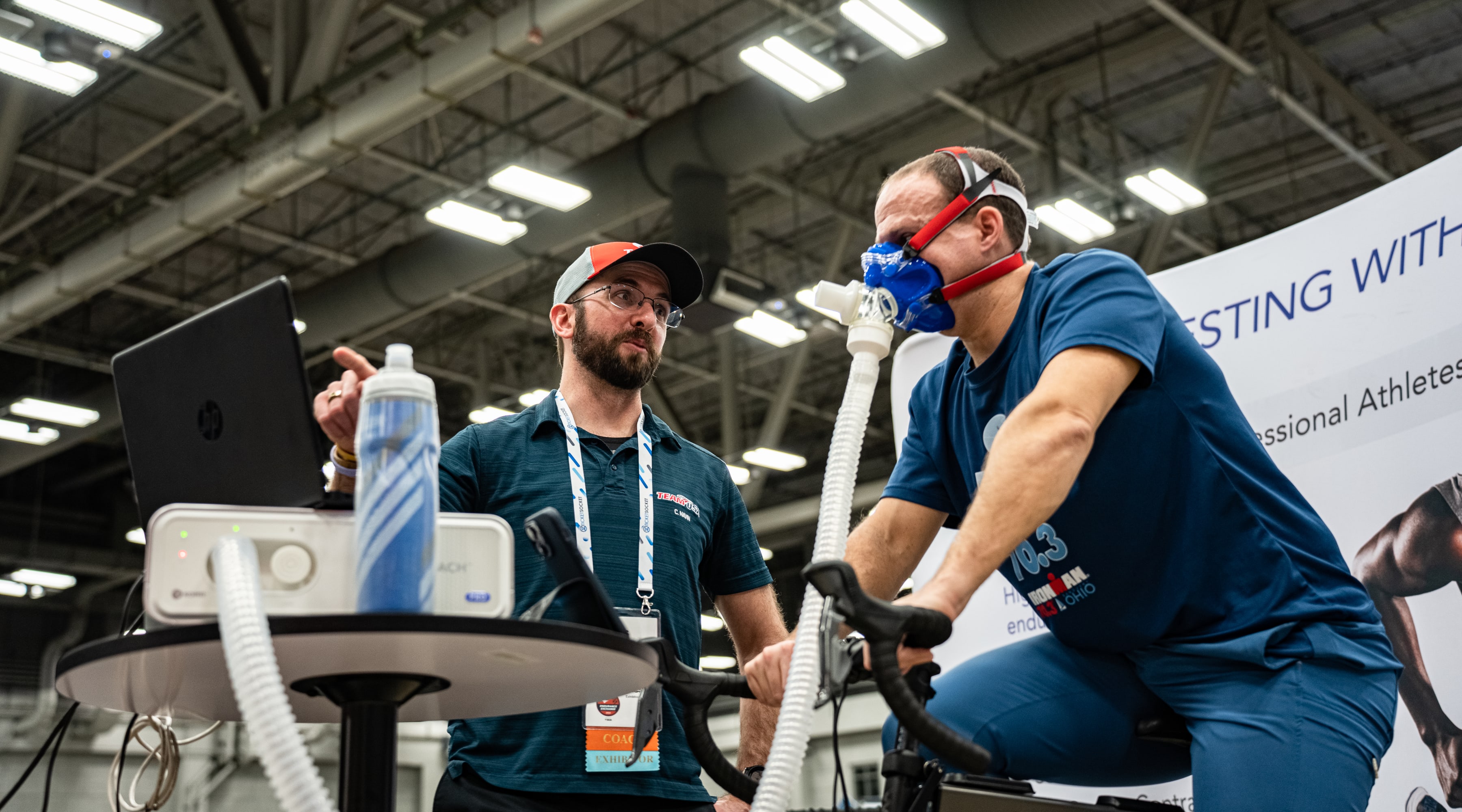 A triathlon coach points at a screen and talks to a cyclist who's riding an indoor trainer with a mask on his face. The mask is attached via a hose to a machine and a computer.