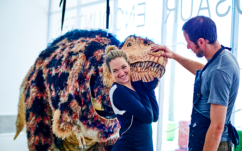 woman stands next to a large-scale dinosaur puppet