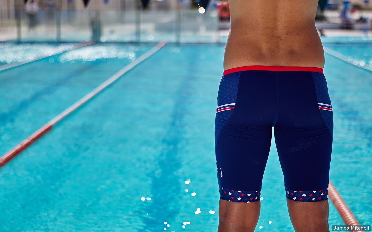 A closeup of a swimmer wearing swim jammers and standing in front of a swimming people with lanes.