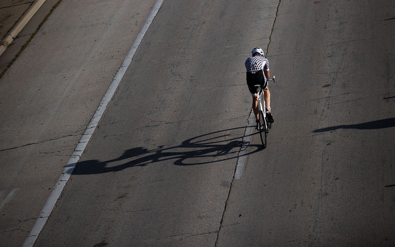 An athlete cycles alone on a closed road. His shadow stretches long to the left of him.