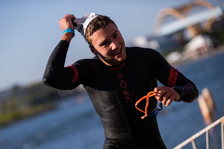 An athlete removes his swim cap and goggles while running out of the water.