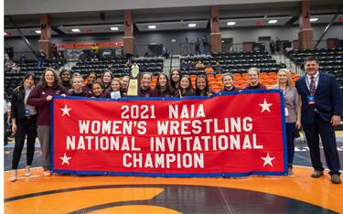 The Campbellsville women's wrestling team poses with its red championship banner after winning the 2021 NAIA Women's Wrestling National Invitational team title.