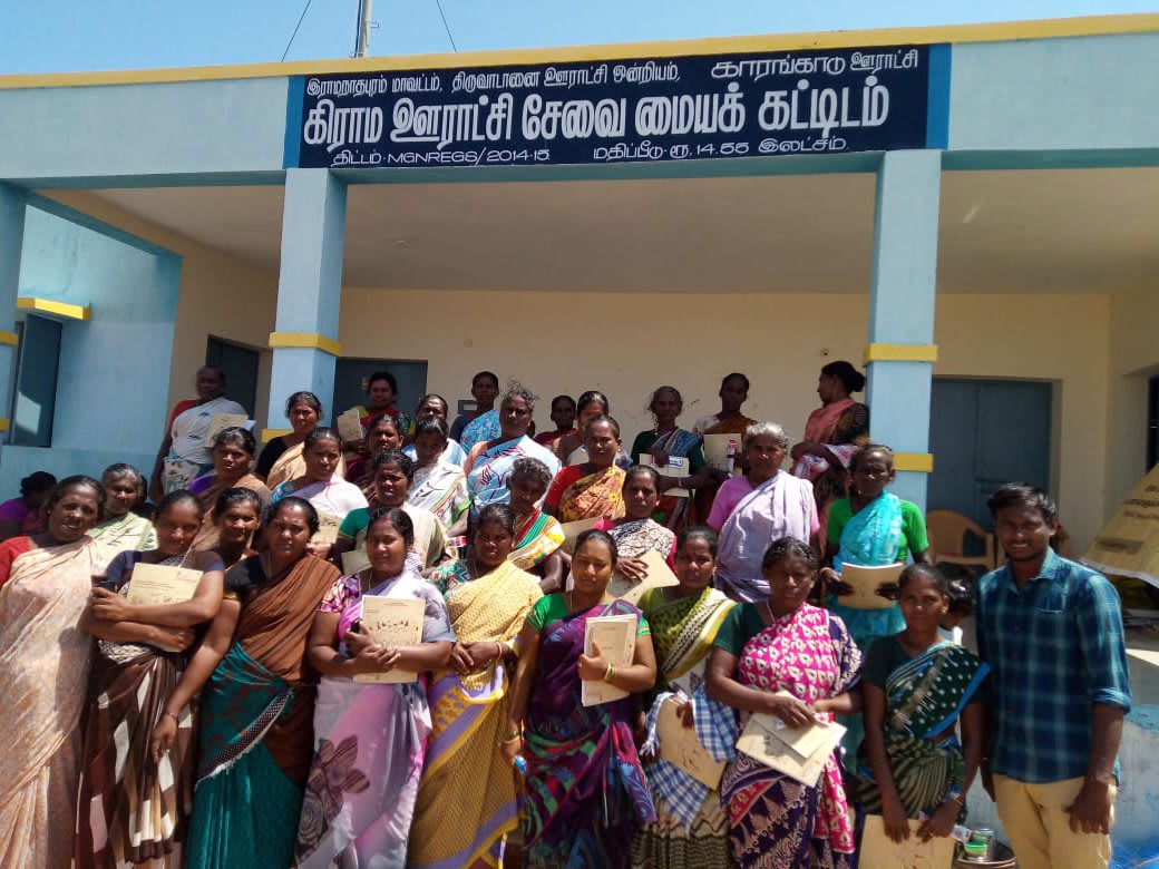 Around thirty women standing in rows outside a blue and white village building, most holding a small printed booklet, a man addressing them from the right