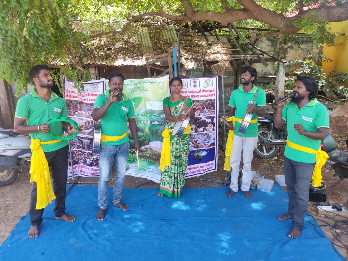 Five performers in green shirts with yellow sashes standing on a blue mat in front of large printed awareness boards, one singing into a microphone