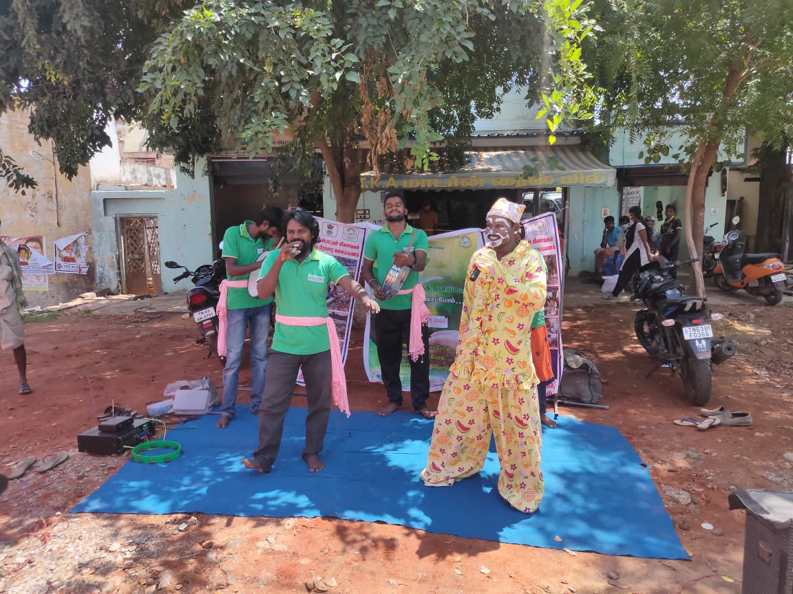 Two performers, one in a patterned costume and cap, acting on a blue mat spread in a village lane while people watch from the roadside