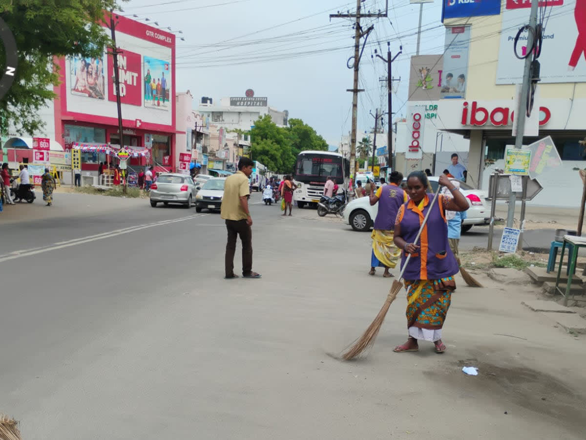 A woman in a sanitation worker tabard sweeping a main road with a long grass broom