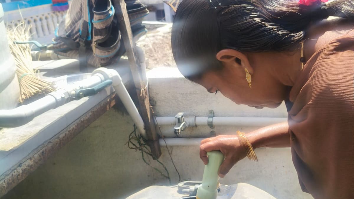 A woman pressing a hand applicator into the open hatch of a stainless-steel water storage tank on a sunlit rooftop terrace, with washing on a line and village rooftops behind her.