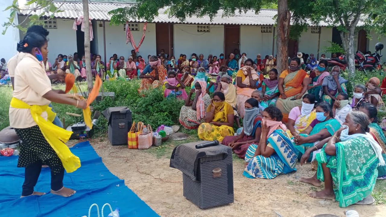A performer in costume addresses a large seated audience of village women under trees at an open-air awareness session