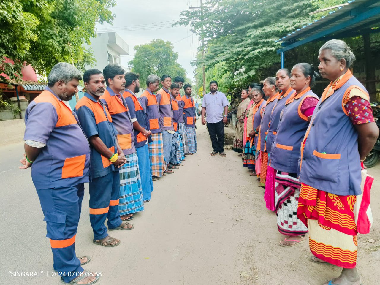 Two facing lines of sanitation workers in grey and orange high-visibility tabards standing along a tree-lined street early in the morning