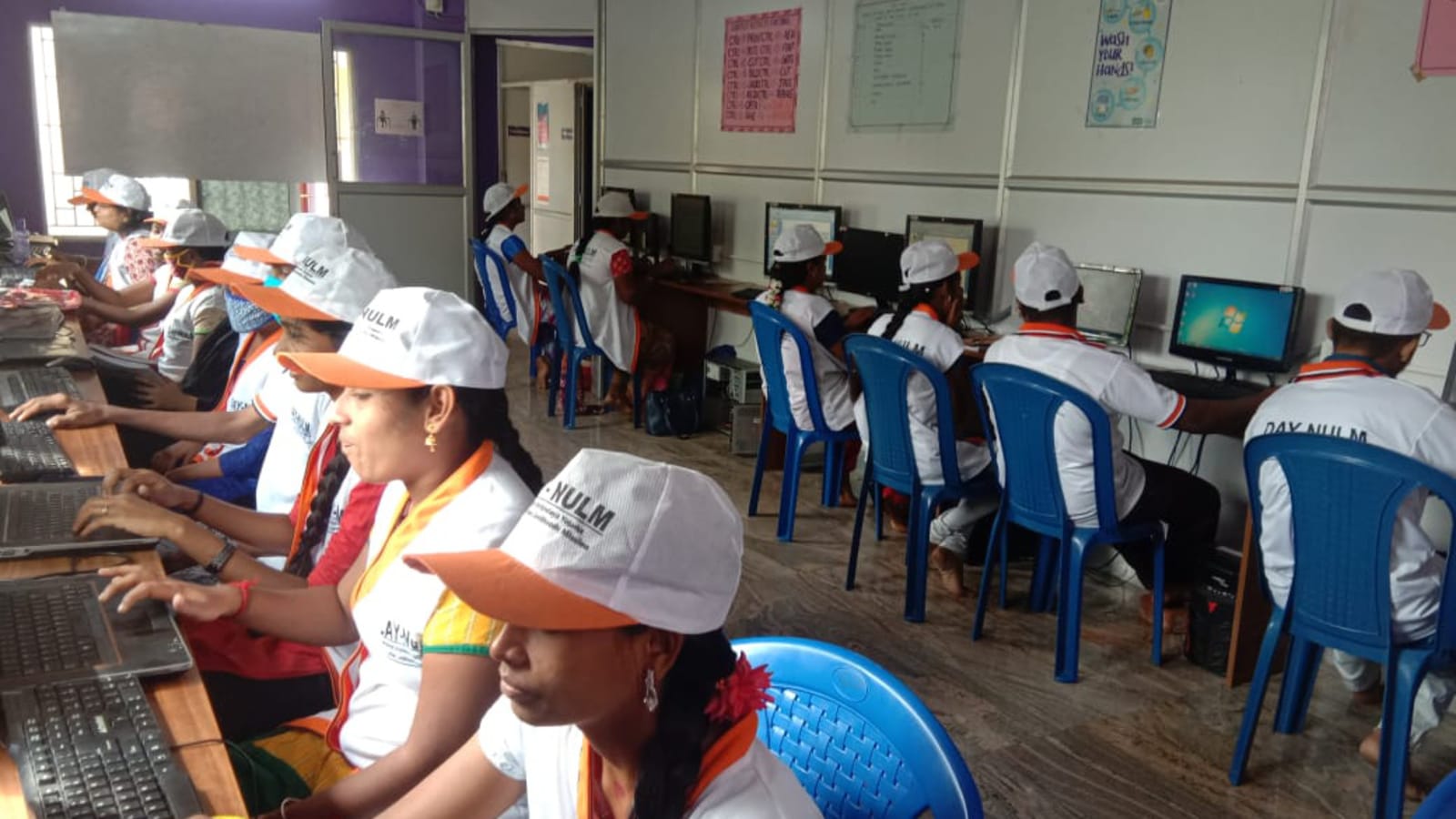 Women in training uniforms typing at laptops along a bench, with more trainees at desktop computers behind them