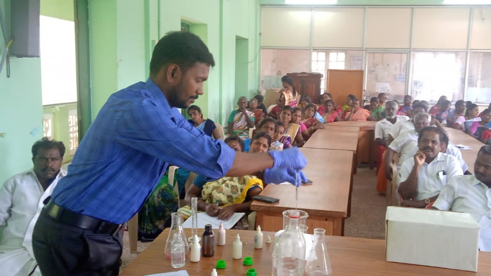 A technician demonstrating a water test with a pipette over sample flasks, with reagent bottles and glassware on the table and community members seated behind