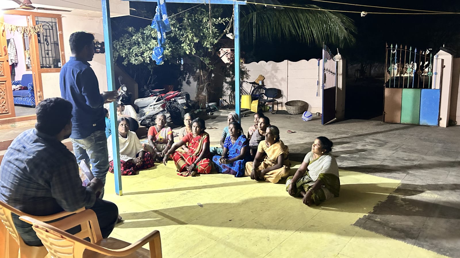 A group of women seated on a courtyard floor at night listening to a man standing with papers