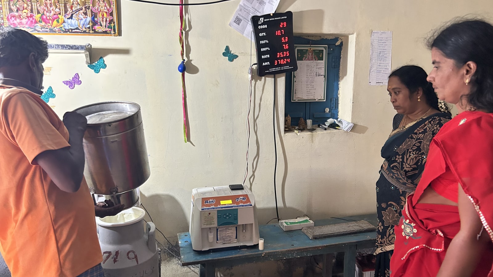 A worker pours milk into a steel can beside a digital milk-testing meter while two women look on inside a collection centre