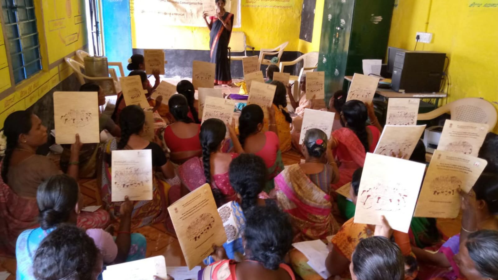 Women seated together indoors holding printed booklets while one woman stands and addresses them