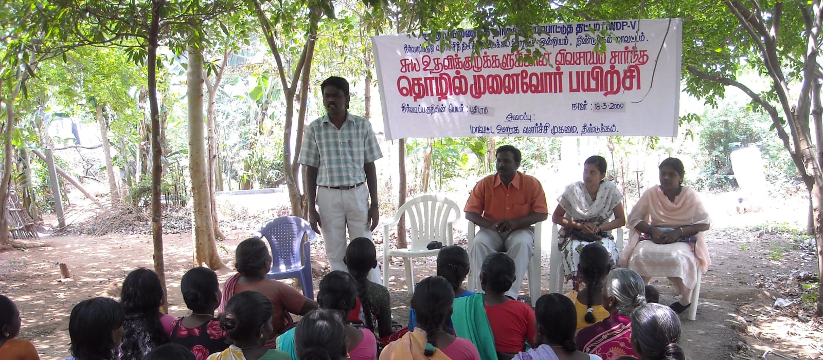 Women seated on the ground at an agriculture-based entrepreneur training session for self-help groups, resource persons seated in front of a Tamil banner