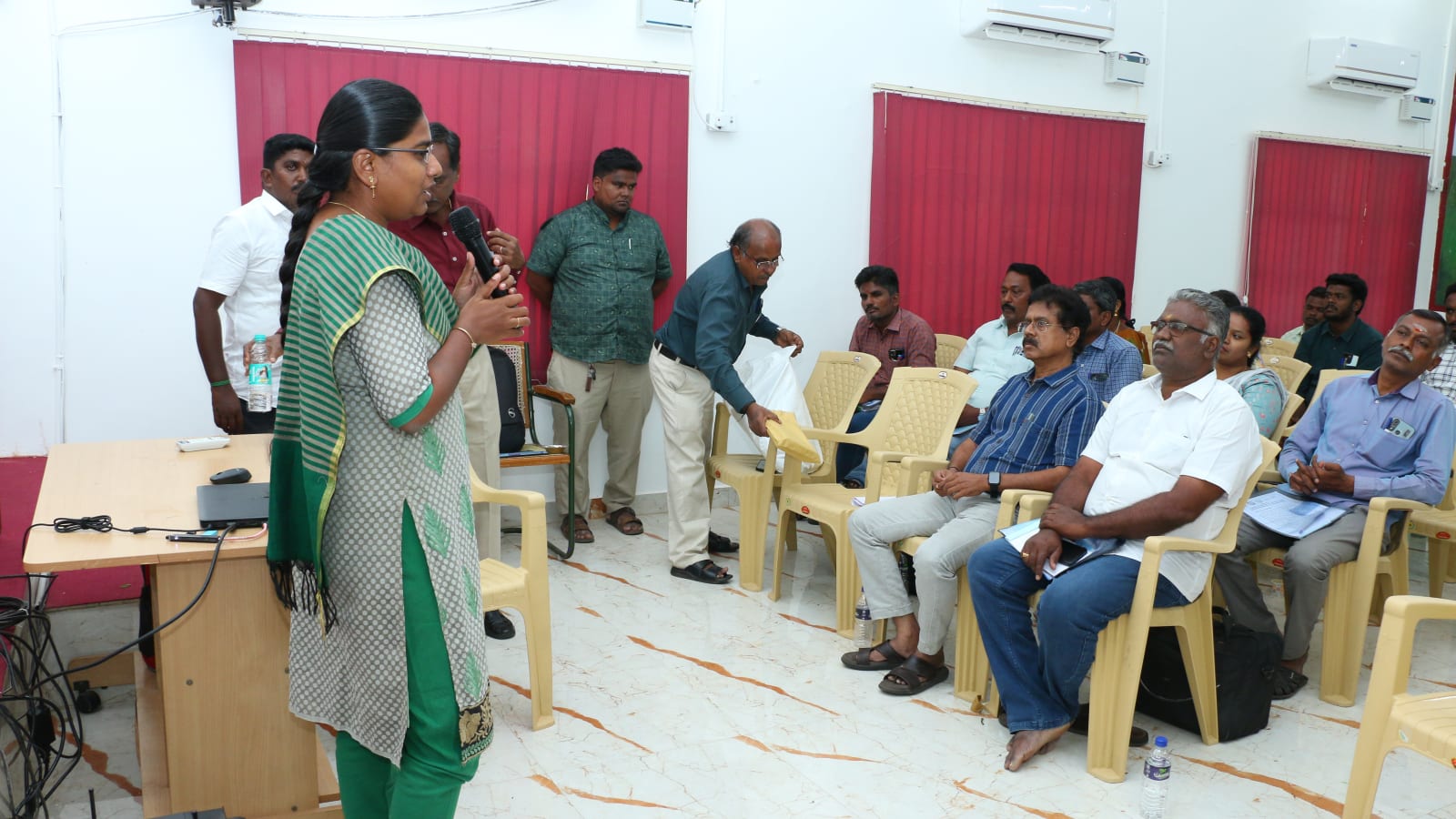 A woman facilitator speaks into a microphone at the front of a training hall while a colleague walks along the rows carrying materials; district staff sit in plastic chairs to the right