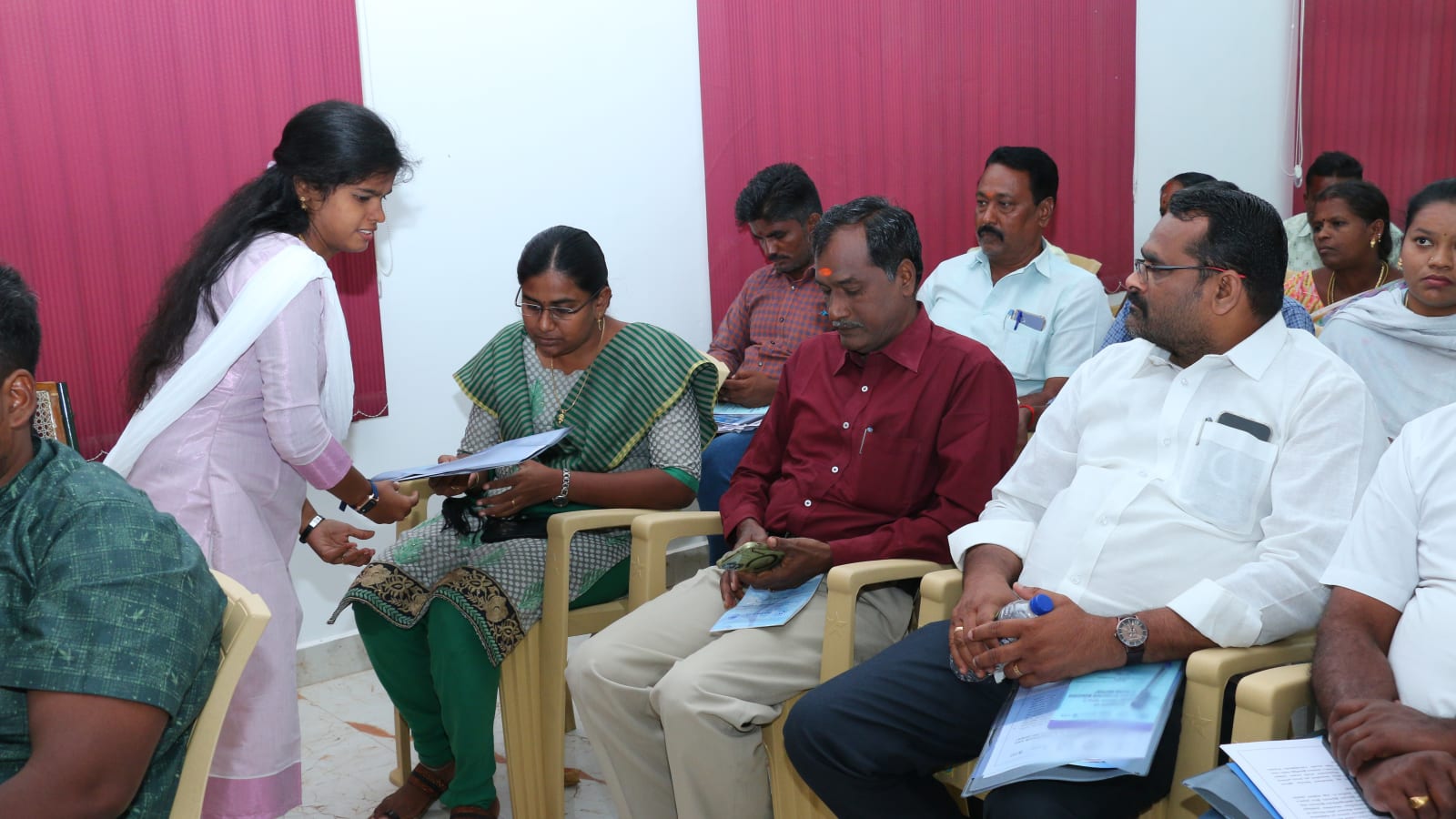 A young woman staff member hands a bound document to a seated woman participant at a training session while other participants hold printed booklets on their laps